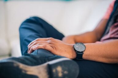 A man sits anxiously on a couch, showcasing his watch, embodying a symptom of depression in men.